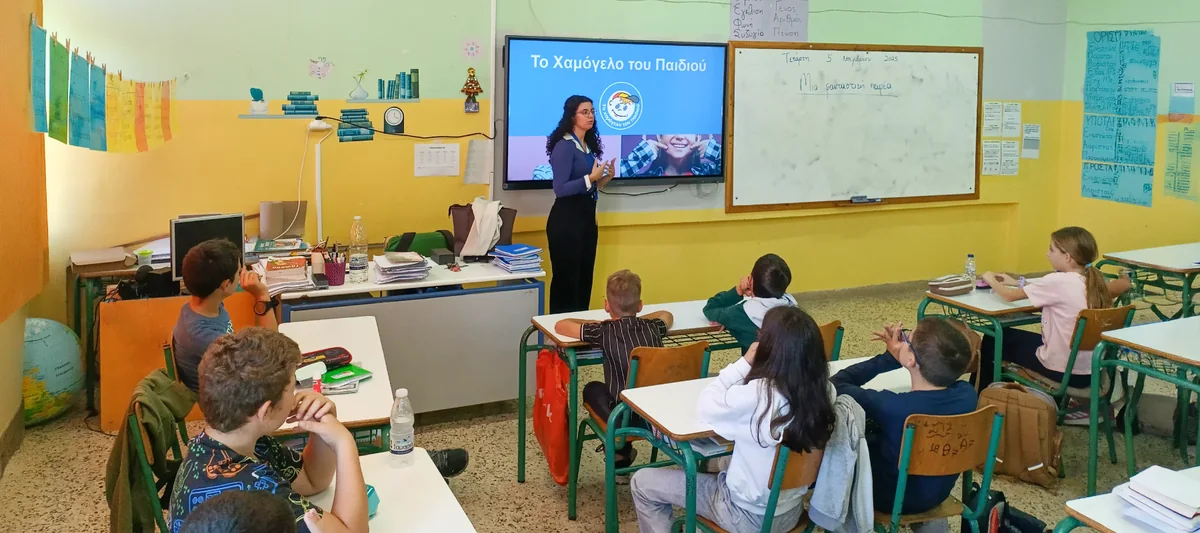 Παρέμβαση του προγράμματος σε σχολική τάξη: ψυχολόγος του Οργανισμού μιλά στα παιδιά μπροστά στην οθόνη με το λογότυπο «Το Χαμόγελο του Παιδιού»