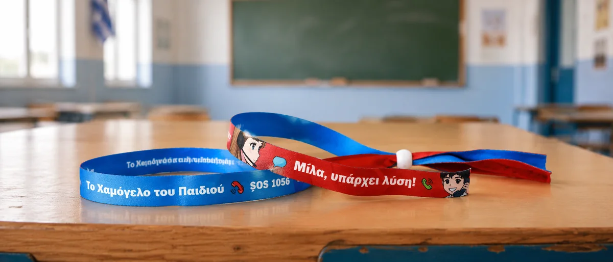 Τα δύο αναμνηστικά βραχιολάκια πάνω σε σχολικό θρανίο: το μπλε «Το Χαμόγελο του Παιδιού — SOS 1056» και το κόκκινο «Μίλα, υπάρχει λύση!»